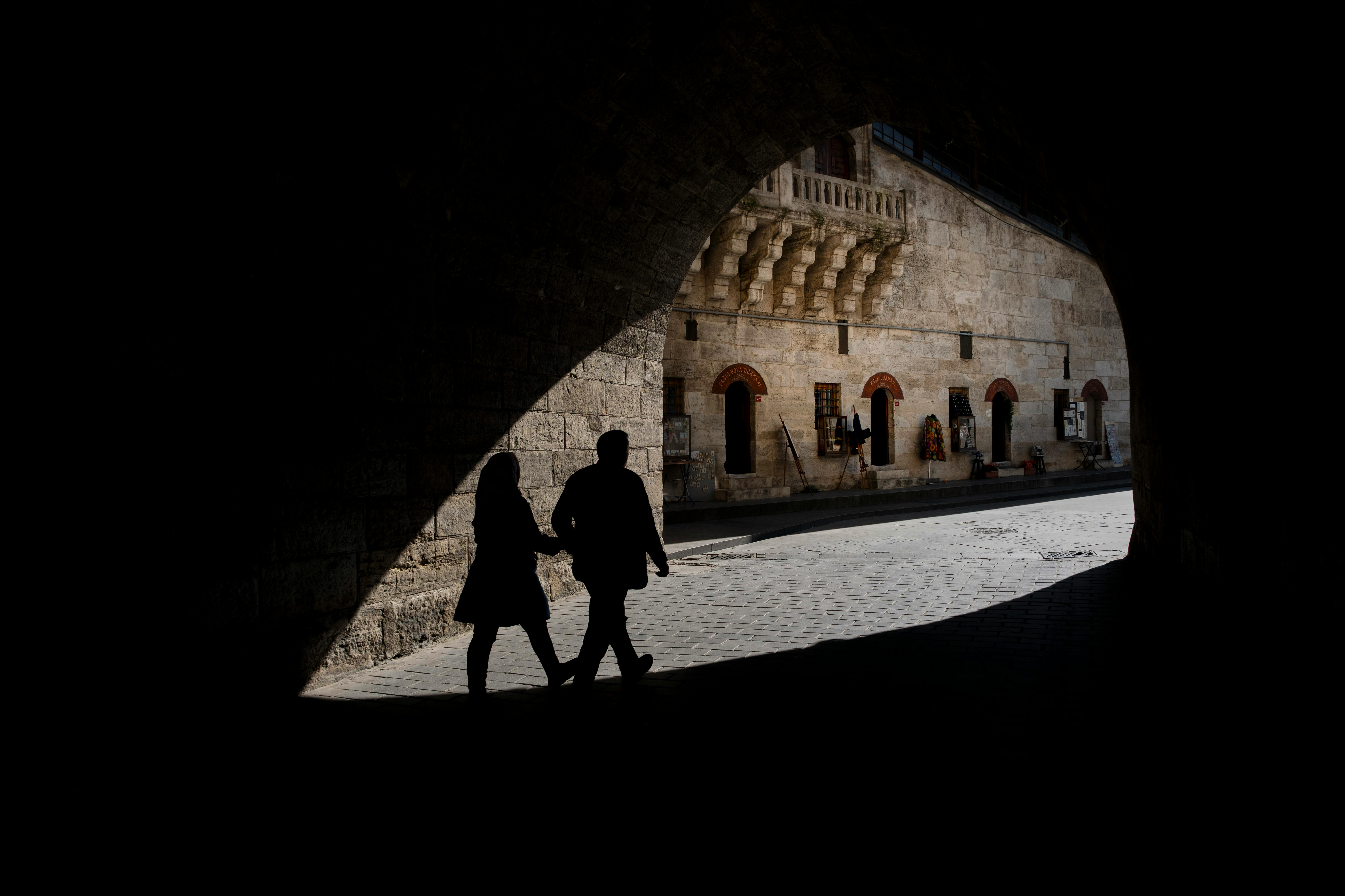 Silhouettes of two people walking under a stone archway in a historic city setting.