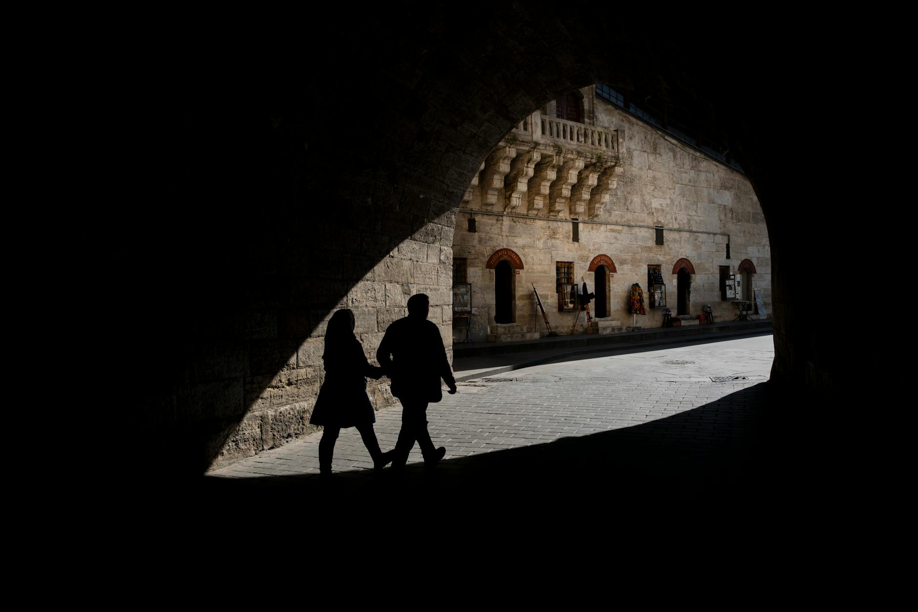 Silhouettes of two people walking under a stone archway in a historic city setting.