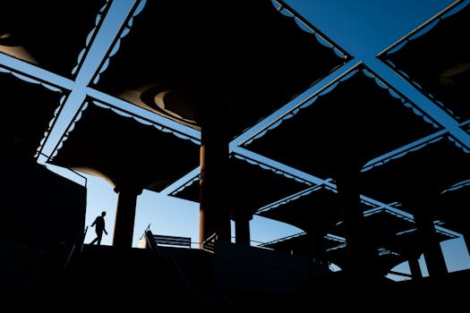 A silhouette of a person walking under a geometric canopy structure with a striking sky background.