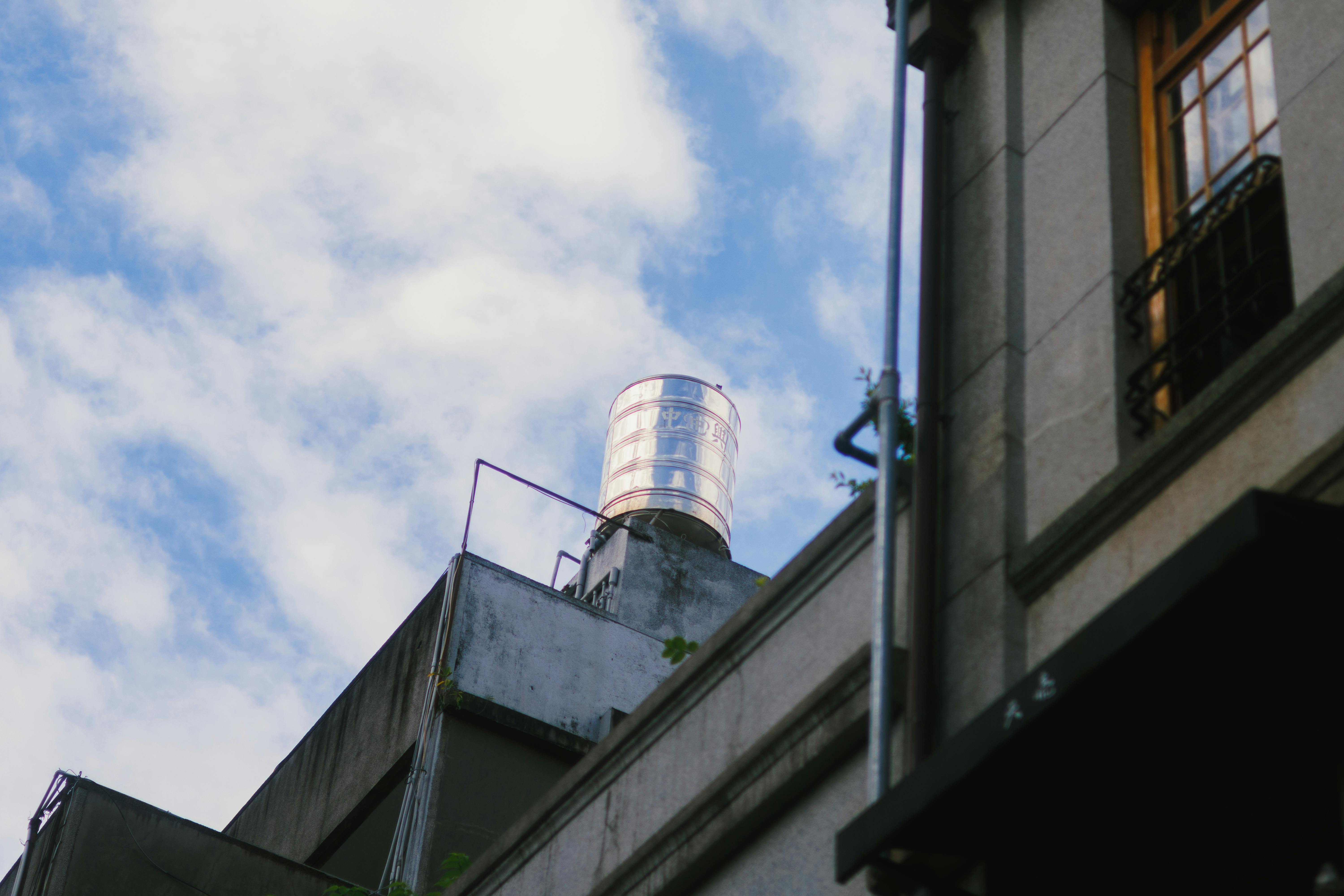 Free A rooftop water tank stands prominently against a vibrant blue sky with scattered clouds. Stock Photo
