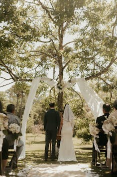A romantic outdoor wedding ceremony with a couple standing under a tree canopy.