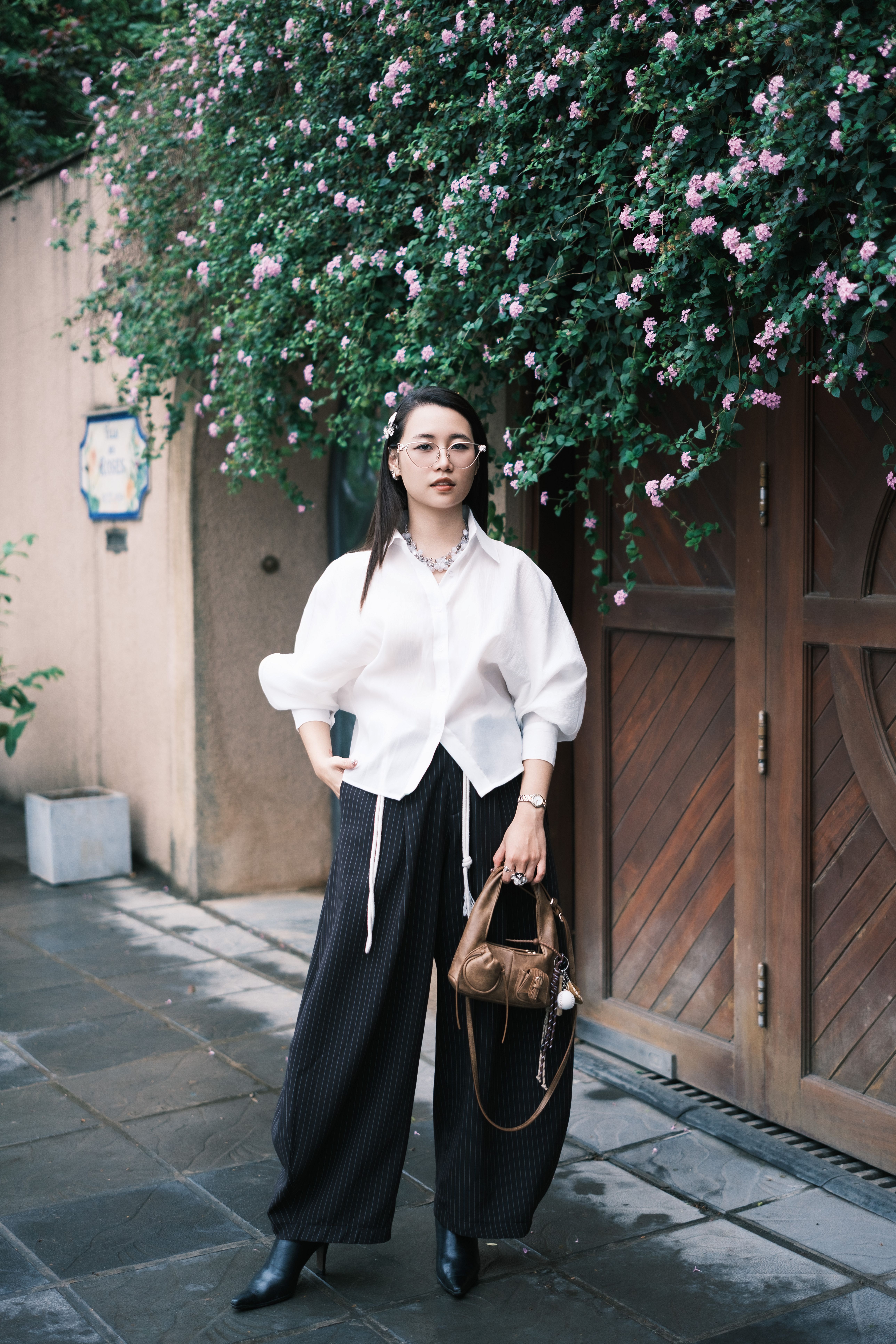 Stylish woman poses under pink flowers in urban setting.