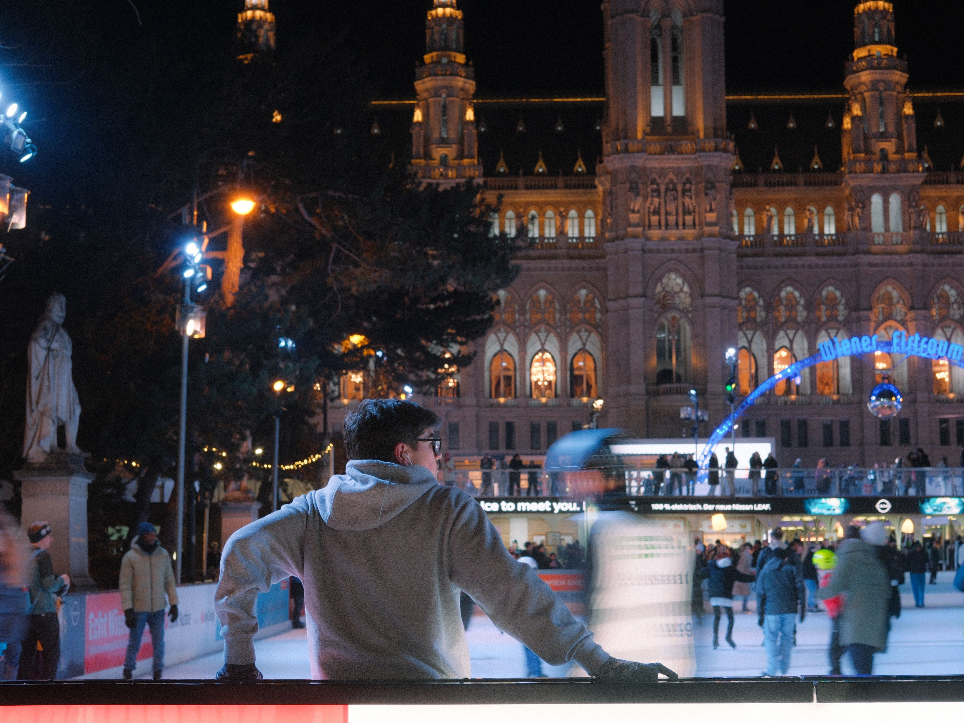 A man in a grey hooded top at the edge of a lit ice rink at night, skaters blurred beyond him and a floodlit town hall with several towers behind