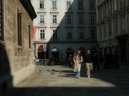 Free Urban street scene with people walking past traditional European architecture, pigeons, and sunlight shadows. Stock Photo