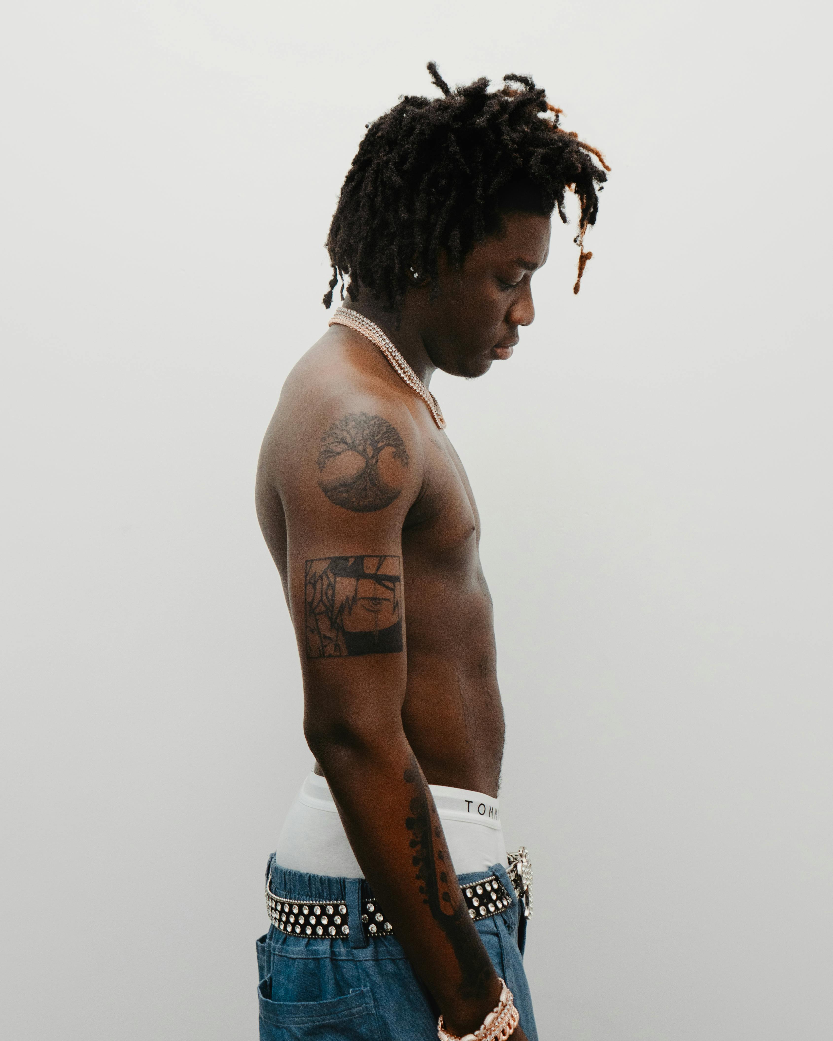 A fashionable young man with tattoos poses in profile against a neutral background.