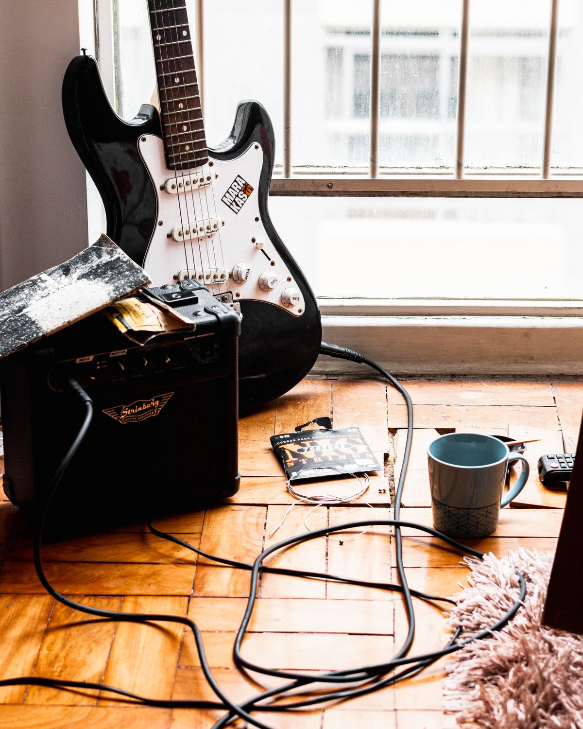 An electric guitar and small amplifier resting on a wooden floor beside a window