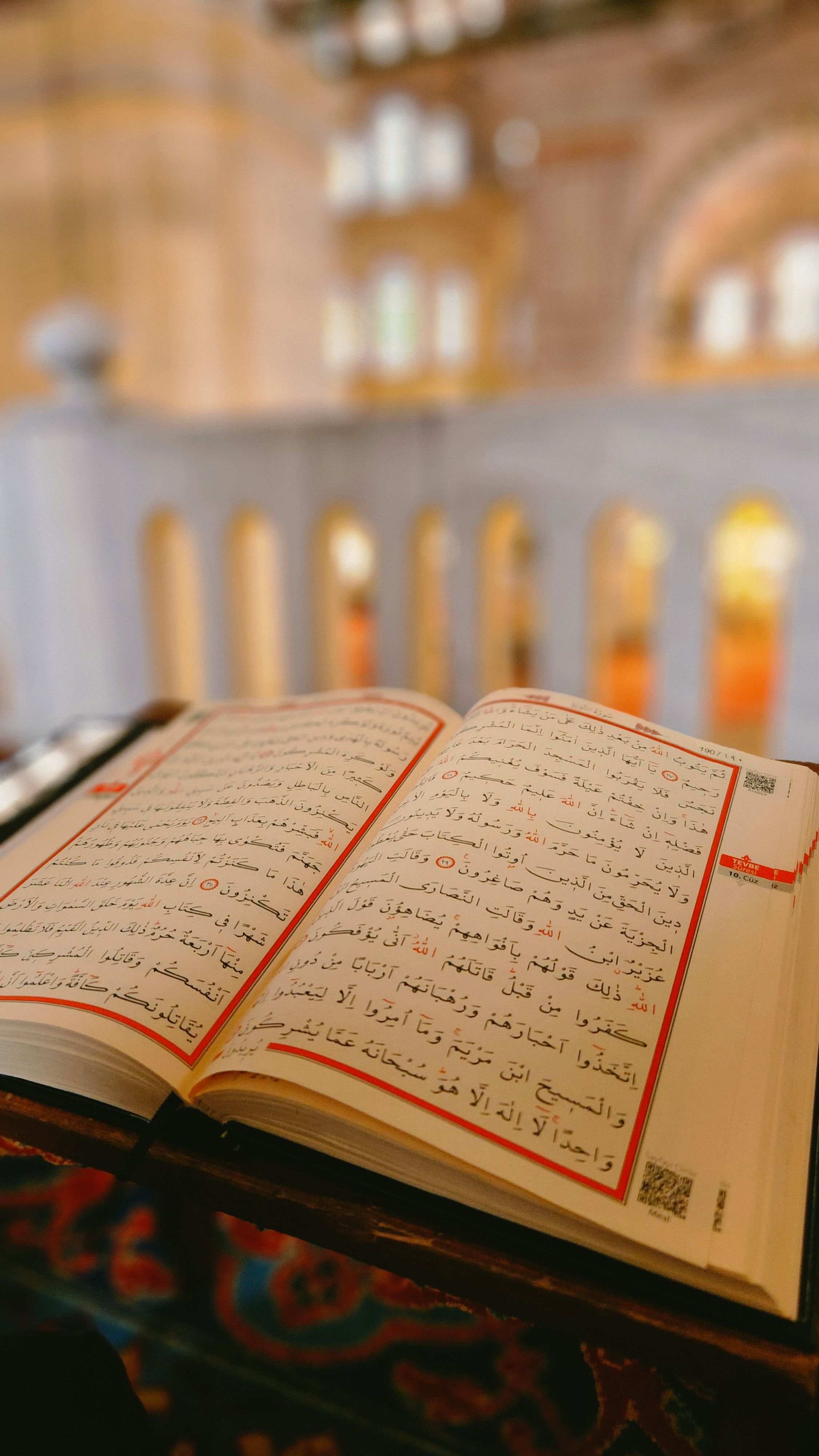 An open Quran on a stand with a blurred mosque interior in the background.