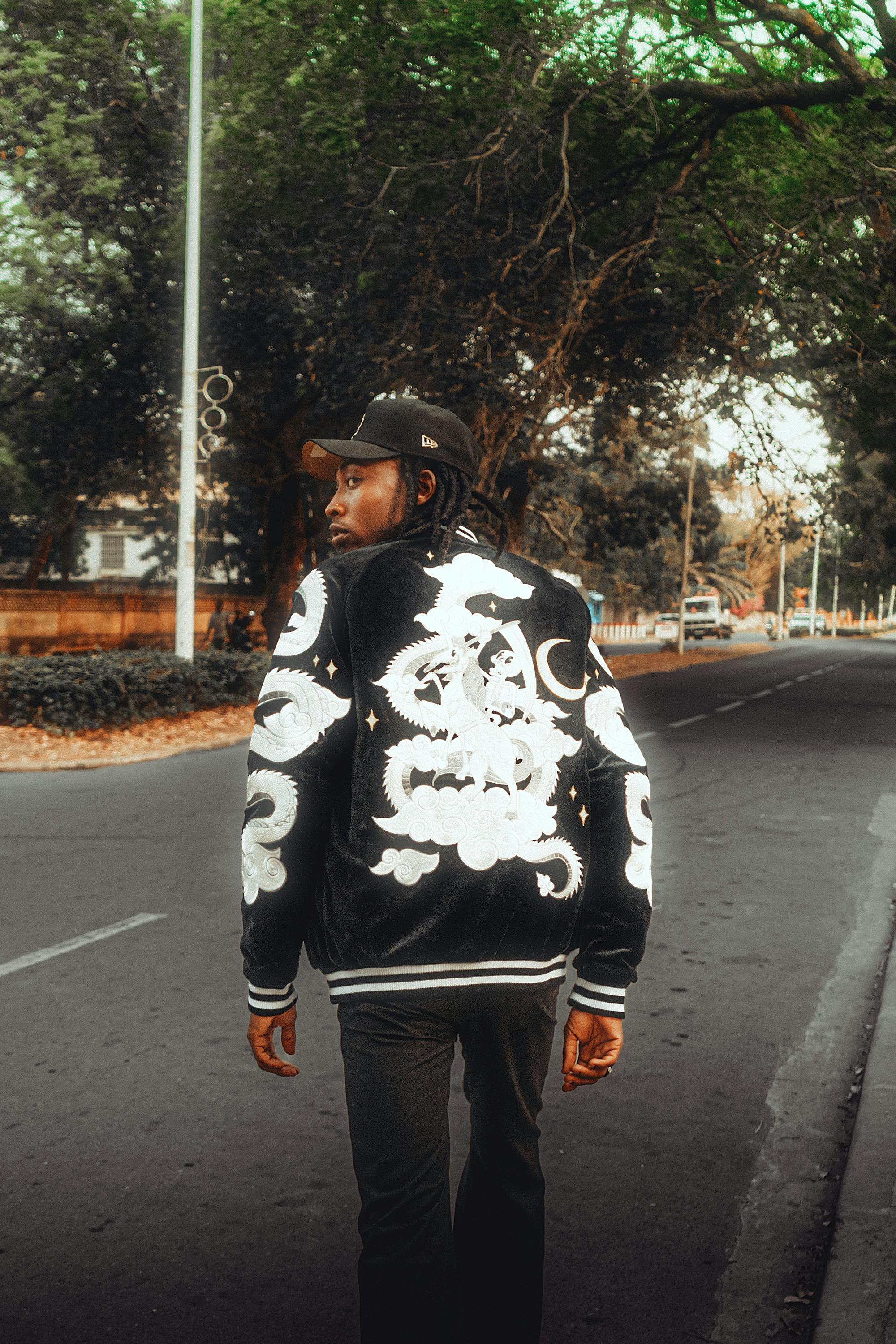 Gratis Un hombre con una chaqueta bordada con un dragón camina por una calle tranquila bordeada de árboles. Foto de stock