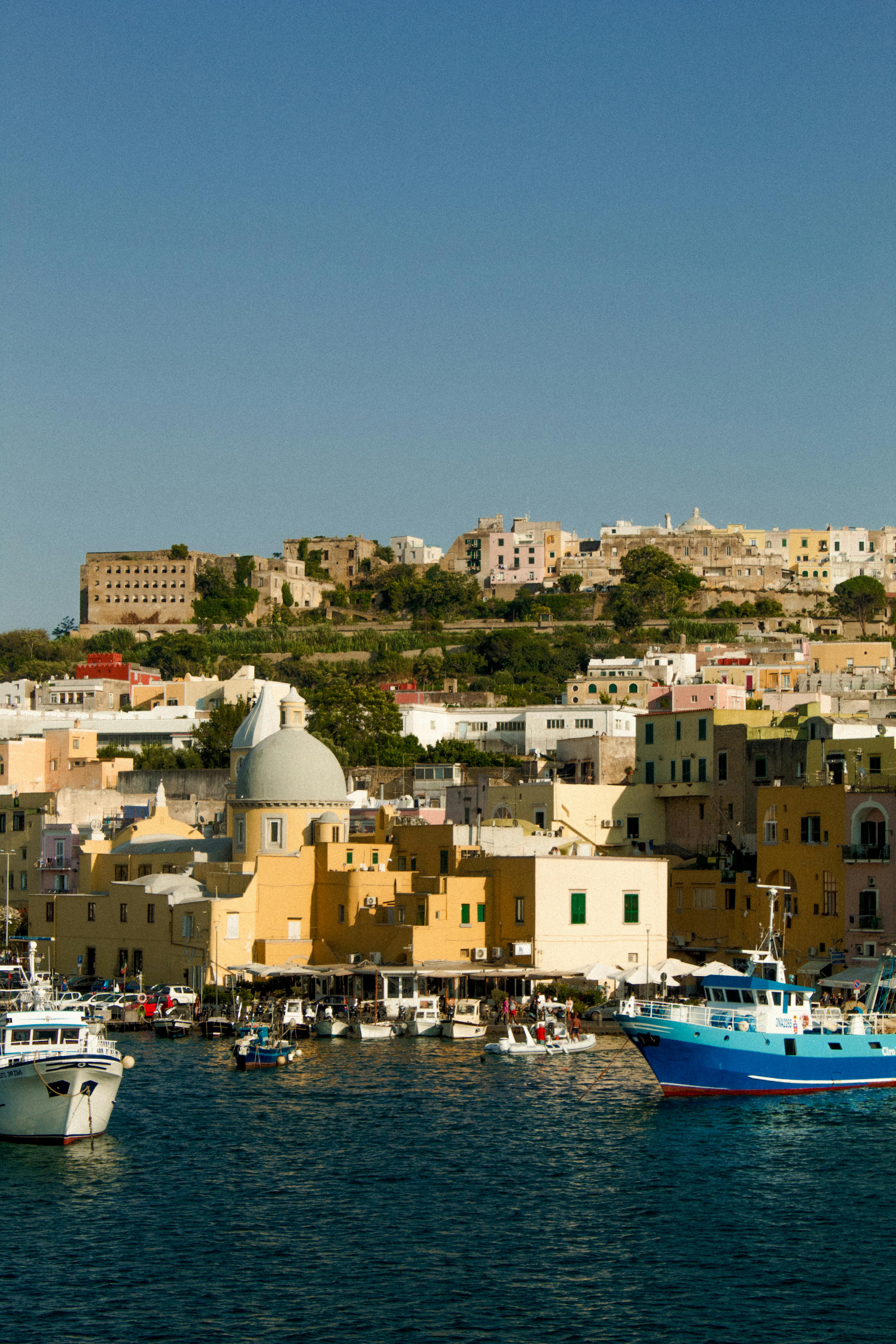 Colorful buildings line the idyllic harbor of Procida Island, Campania, Italy, under a clear blue sky.