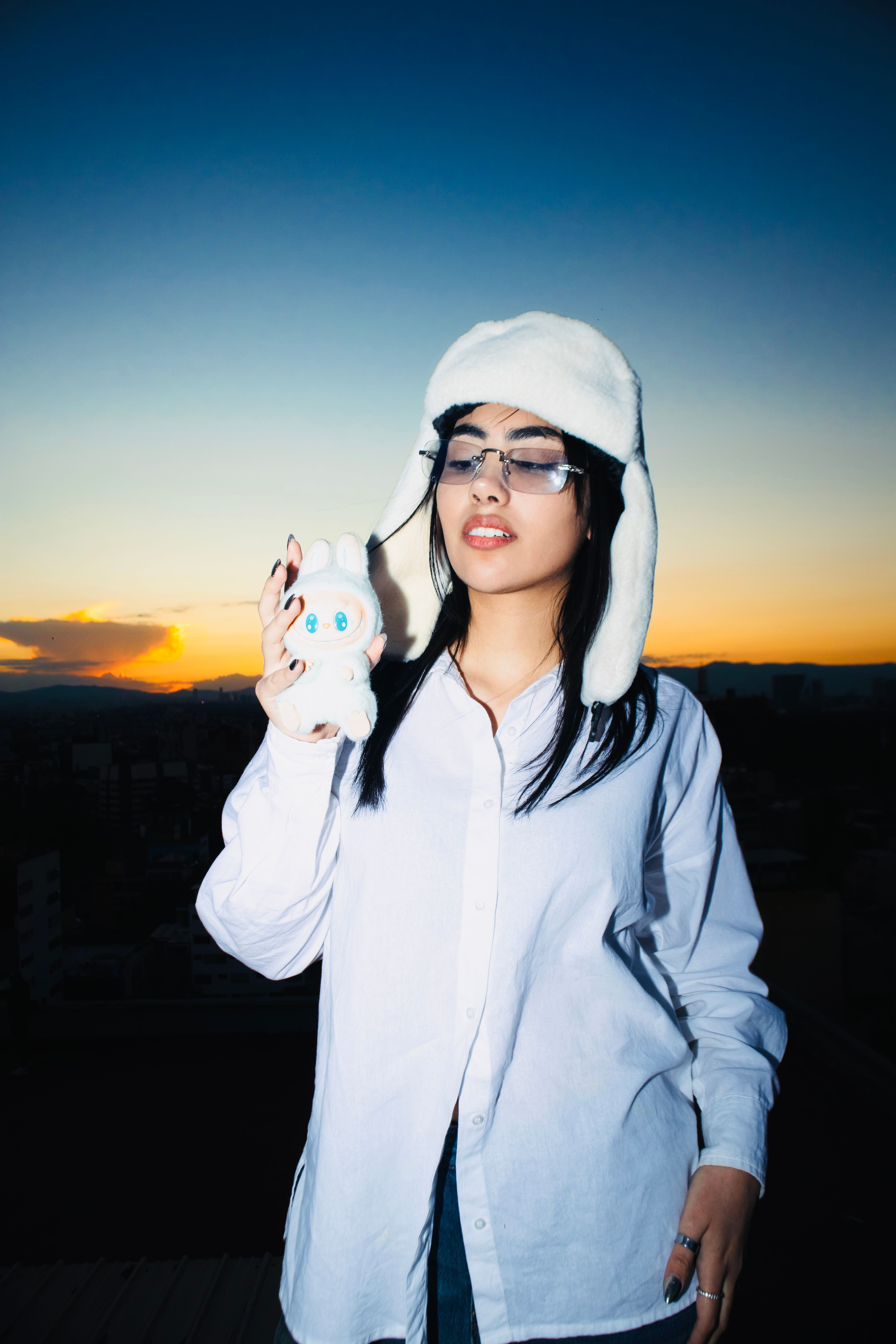 Stylish young woman in white hat and shirt holding a plush toy at sunset in Mexico City.
