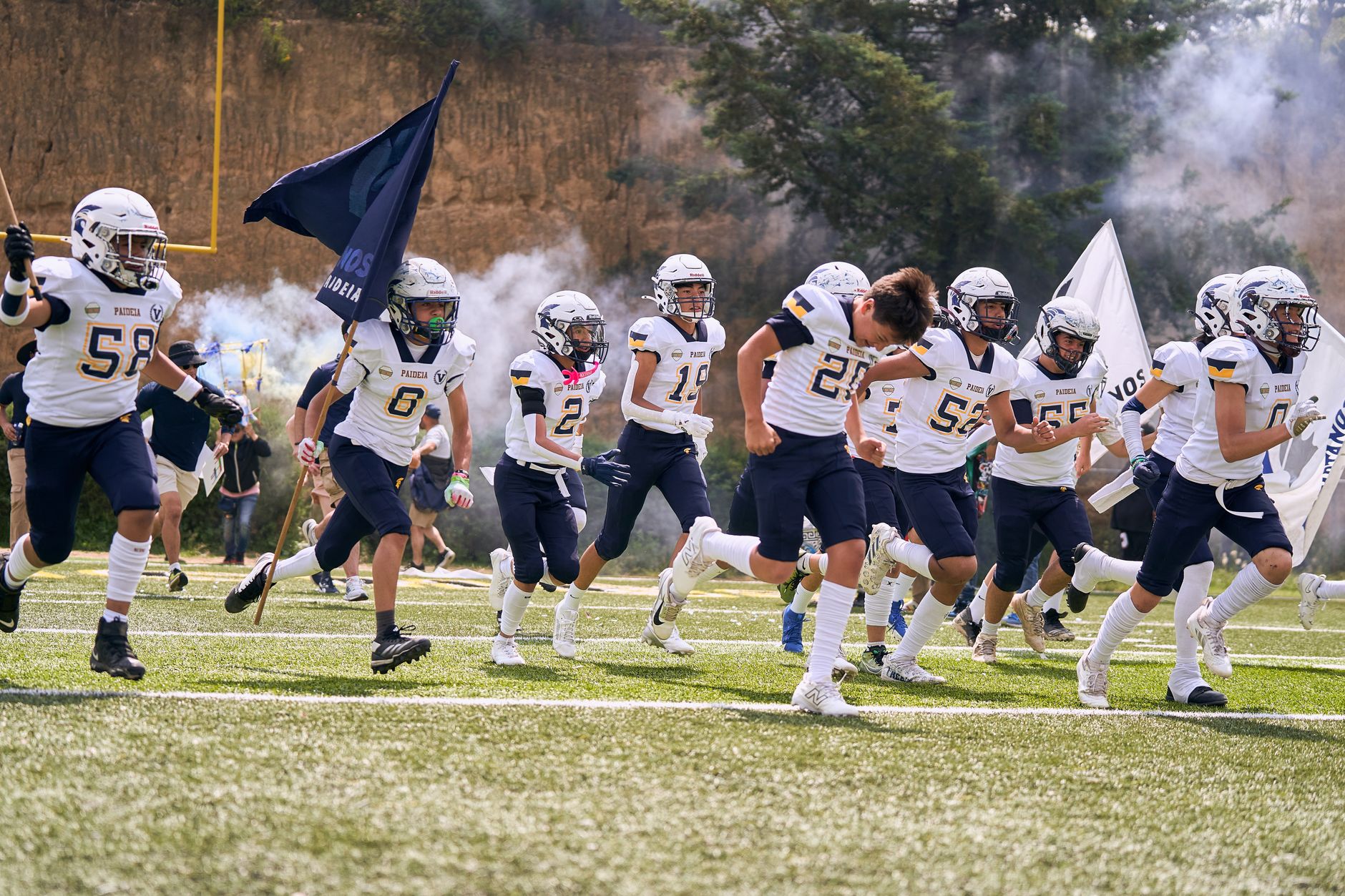 A high school football team energetically runs onto the field with flags under clear skies.