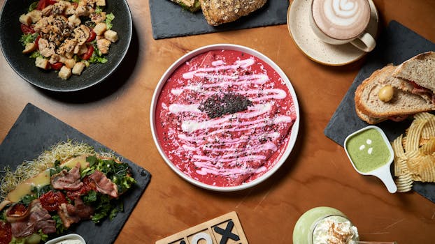 Flat lay of various gourmet dishes including pasta, sandwiches, and latte on a wooden table.