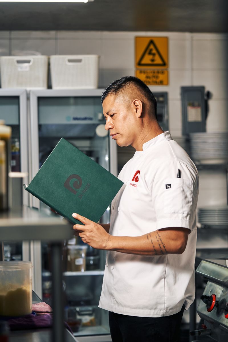 Chef in white uniform examines a recipe book in a professional kitchen setting.