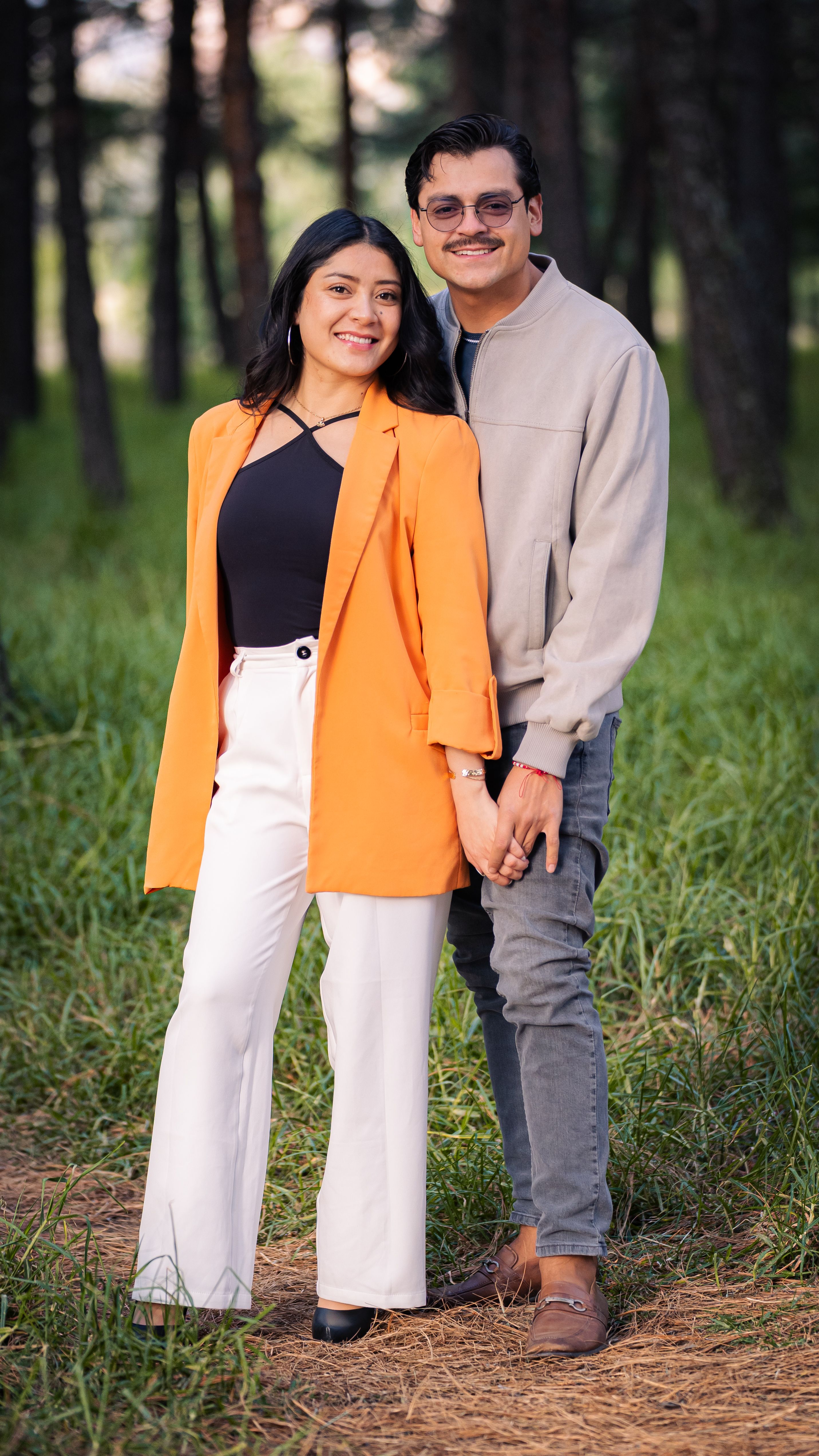 A cheerful couple poses together in a lush green forest, smiling warmly at the camera.