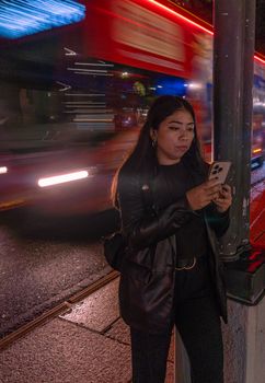 Young woman leaning against a post, using a smartphone in a vivid city scene with a bus speeding by at night.