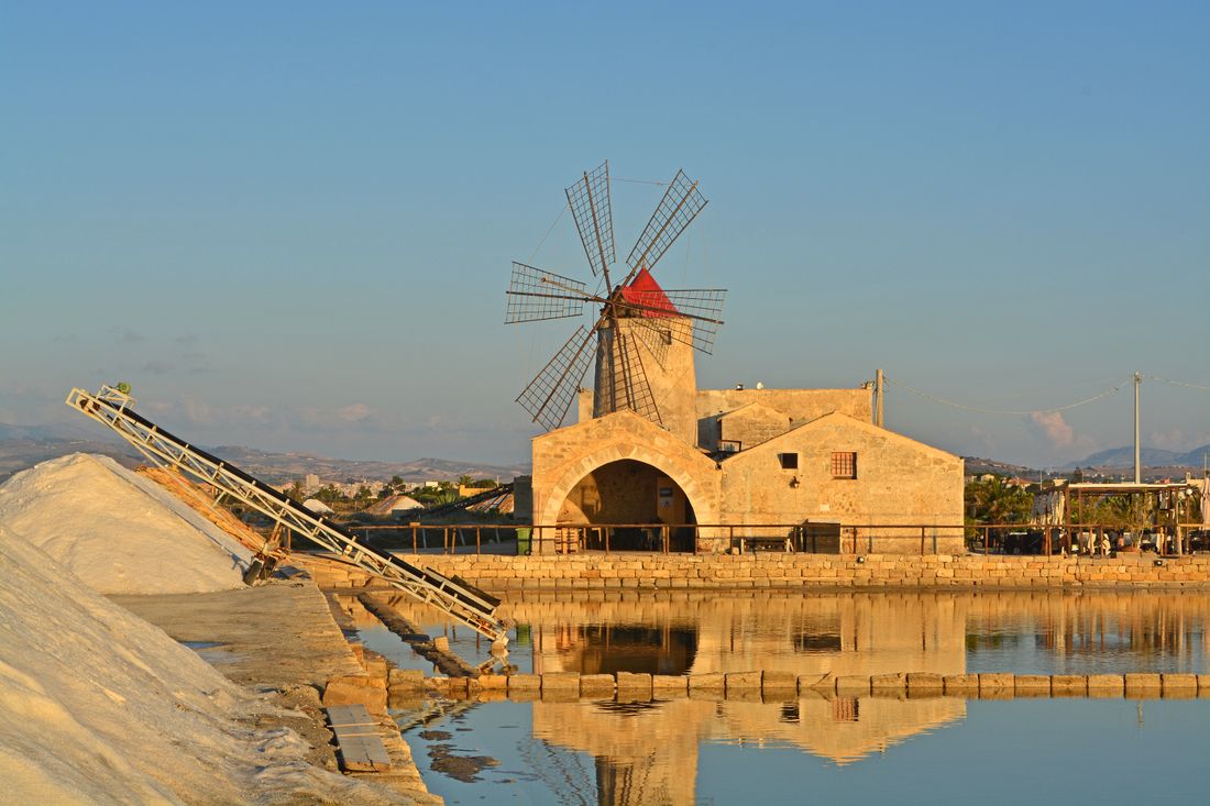 A stone salt mill with a red-capped windmill reflected in a salt pan at sunset, a white salt heap beside it