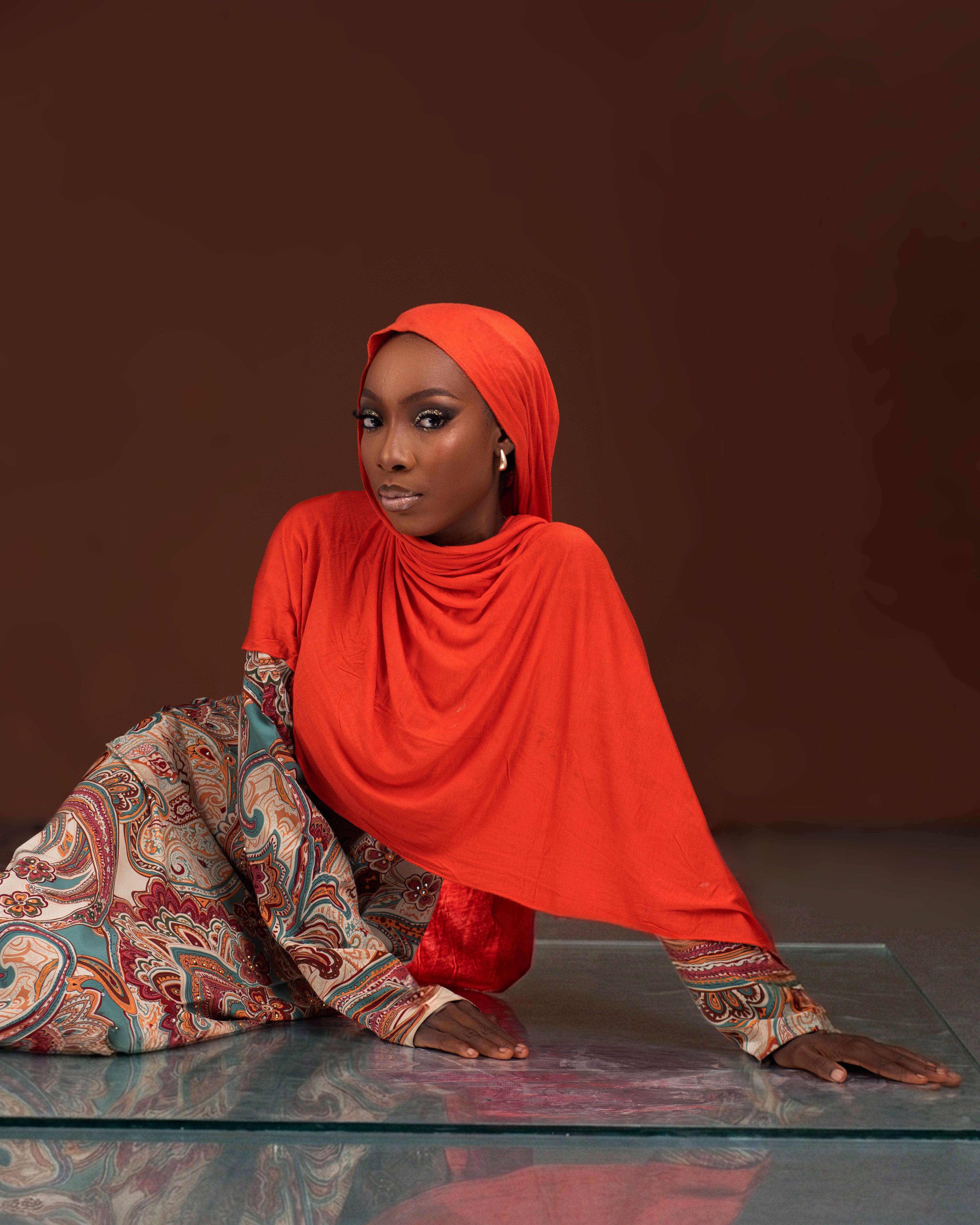 Woman in vibrant red hijab with paisley dress posing against a brown background.