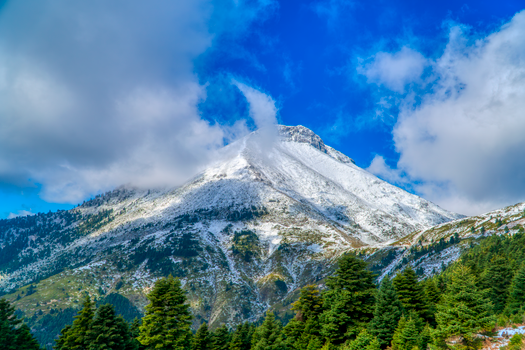 A stunning snow-capped mountain peak in Greece with surrounding lush greenery under a vibrant blue sky.