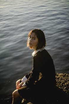 从上方看去，一位身穿黑衣的悲伤年轻女子正坐在河岸的石头上，沐浴着阳光