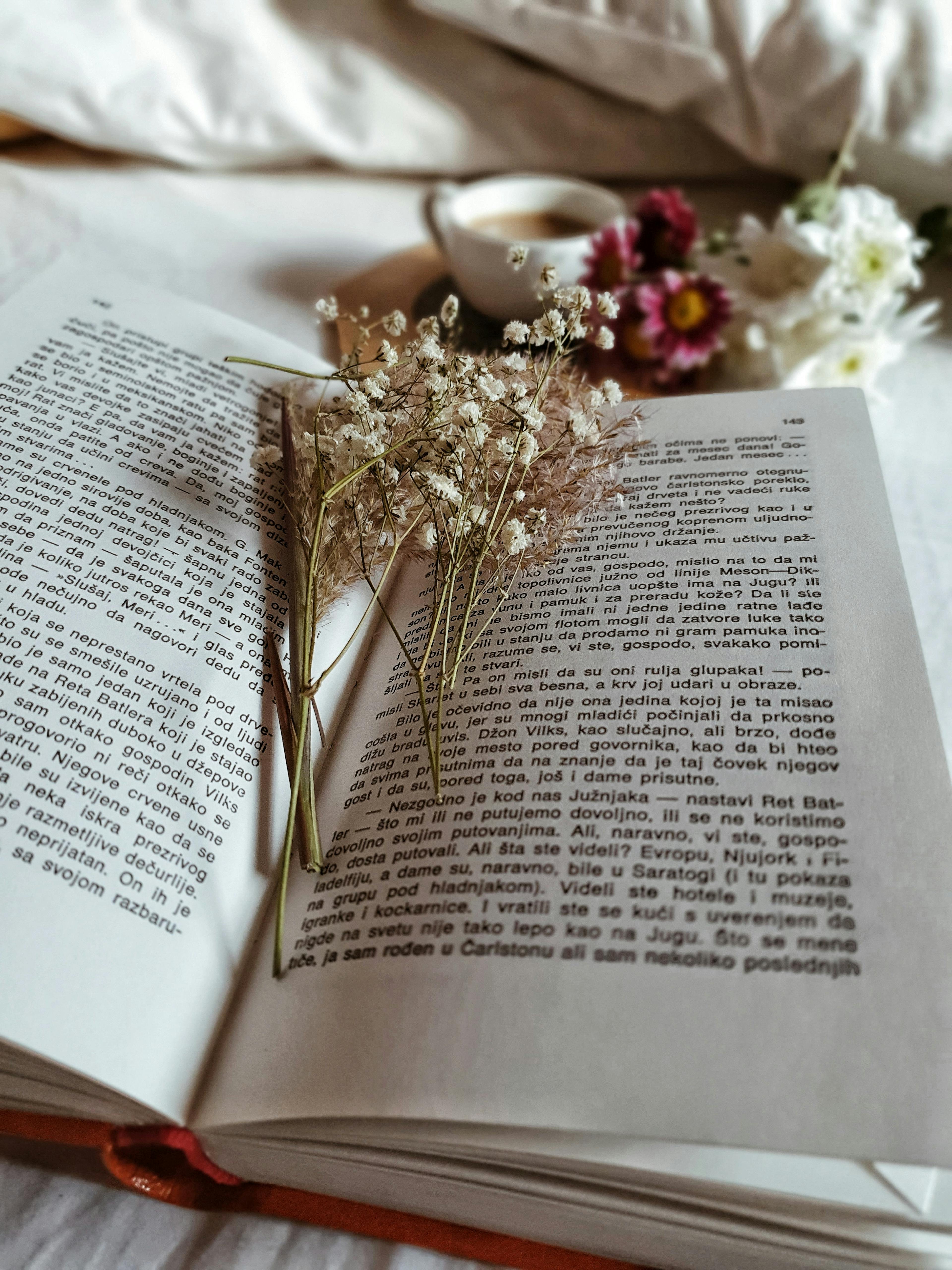 From above of opened book with dried flowers and cup of coffee with fresh flowers in bed at morning time