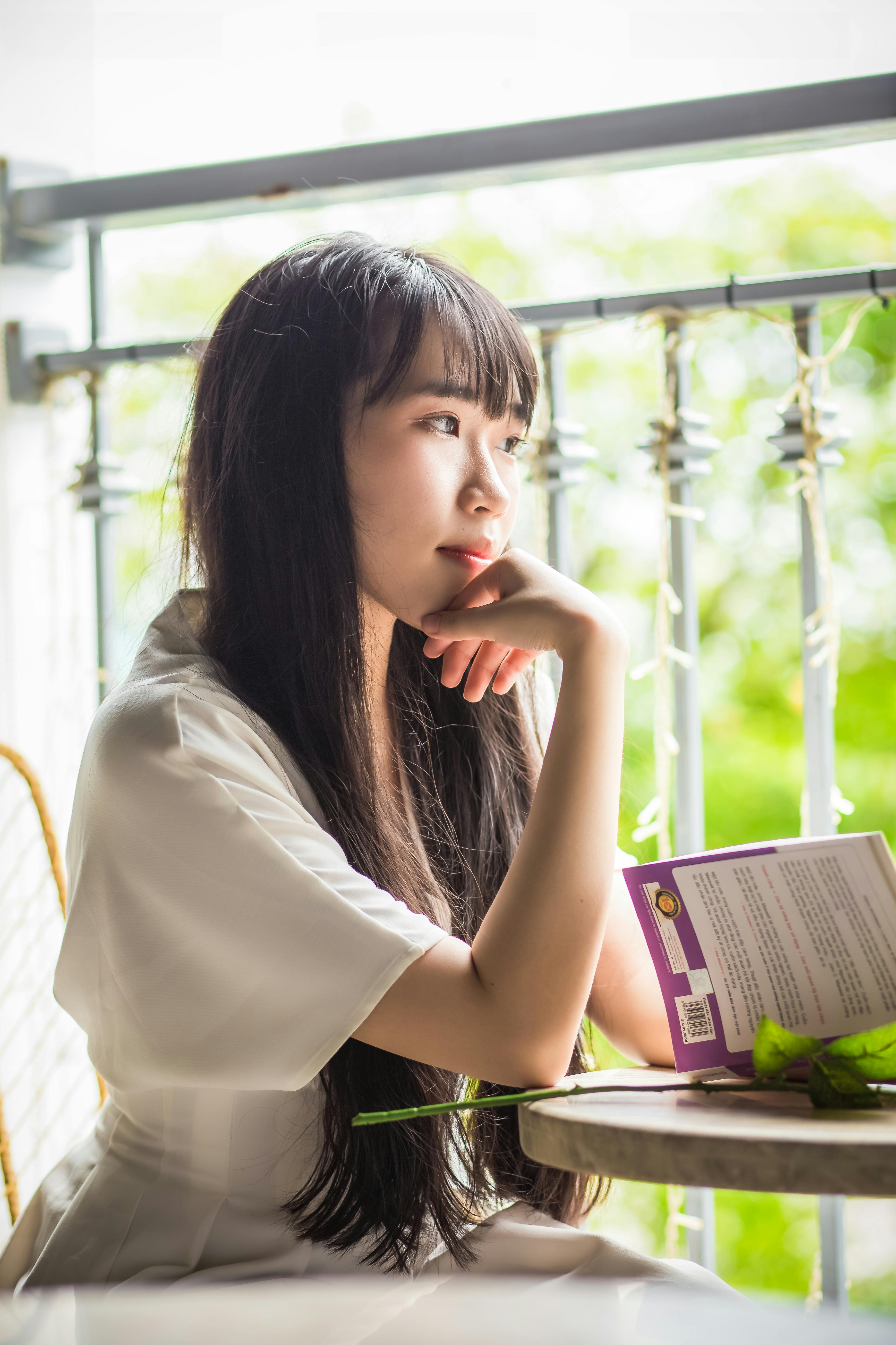 一名东亚妇女身穿白色连衣裙，坐在阳光明媚的窗前看书