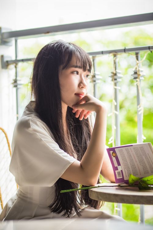 Free An East Asian woman in a white dress sits indoors reading a book by a sunny window. Stock Photo