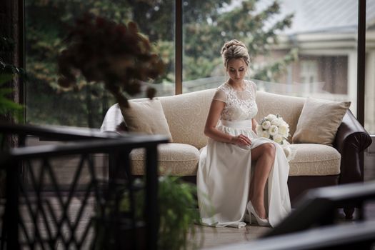 Bride sits gracefully on sofa, holding bouquet, in bright, serene setting.