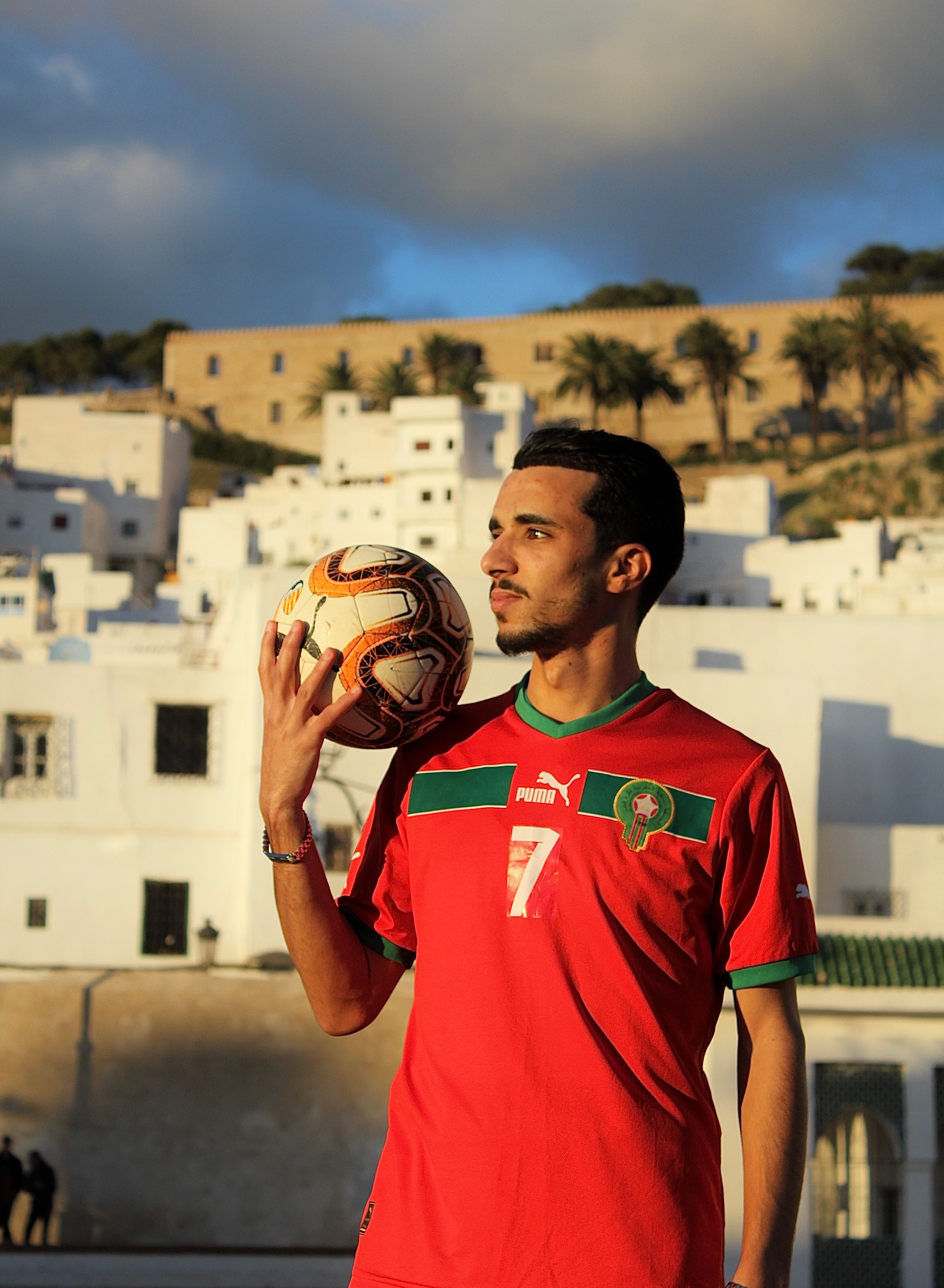 Young Moroccan footballer in Tétouan cityscape holding soccer ball at sunset.