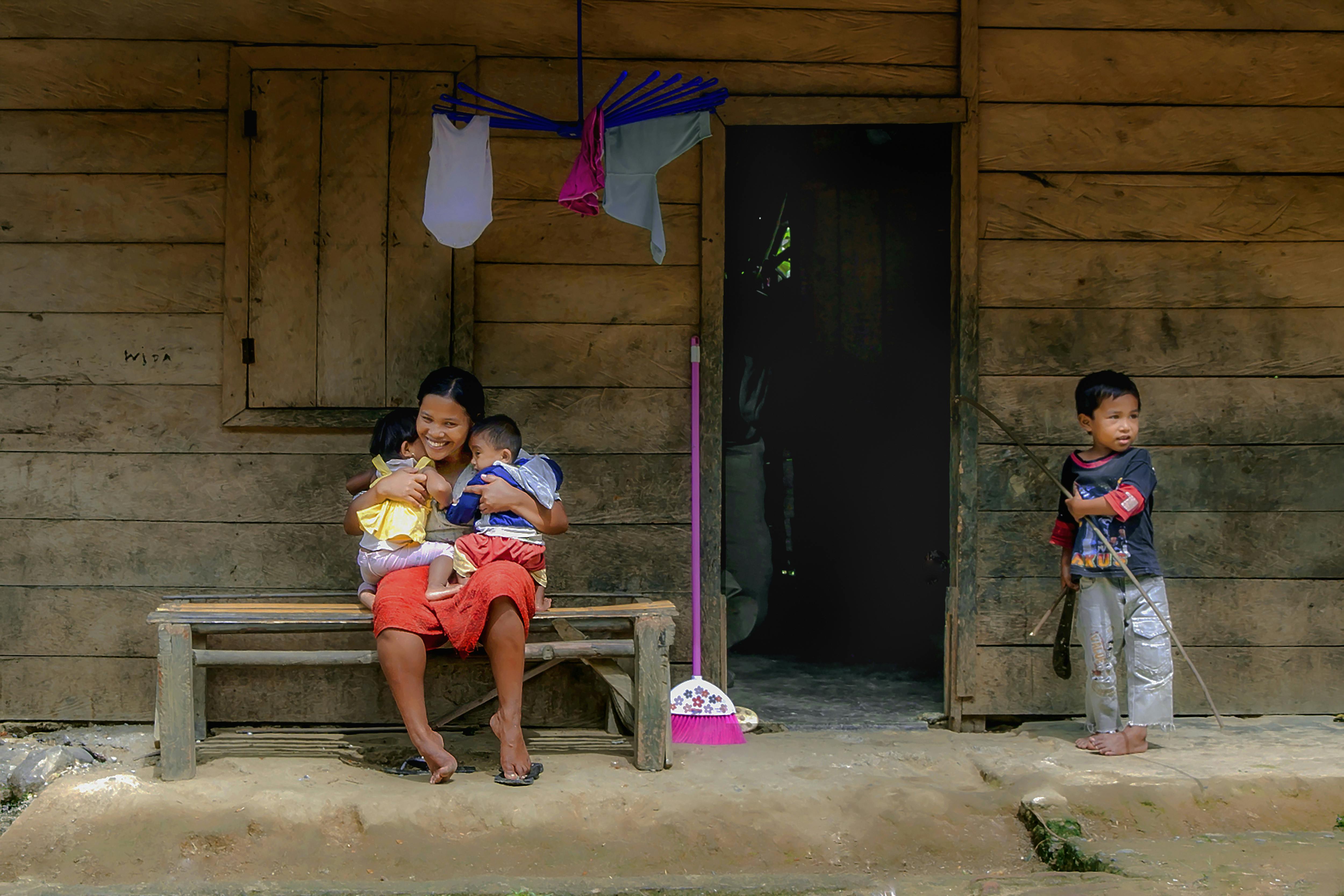 A joyful Indonesian mother and children relaxing outside a rustic wooden home, capturing family life.