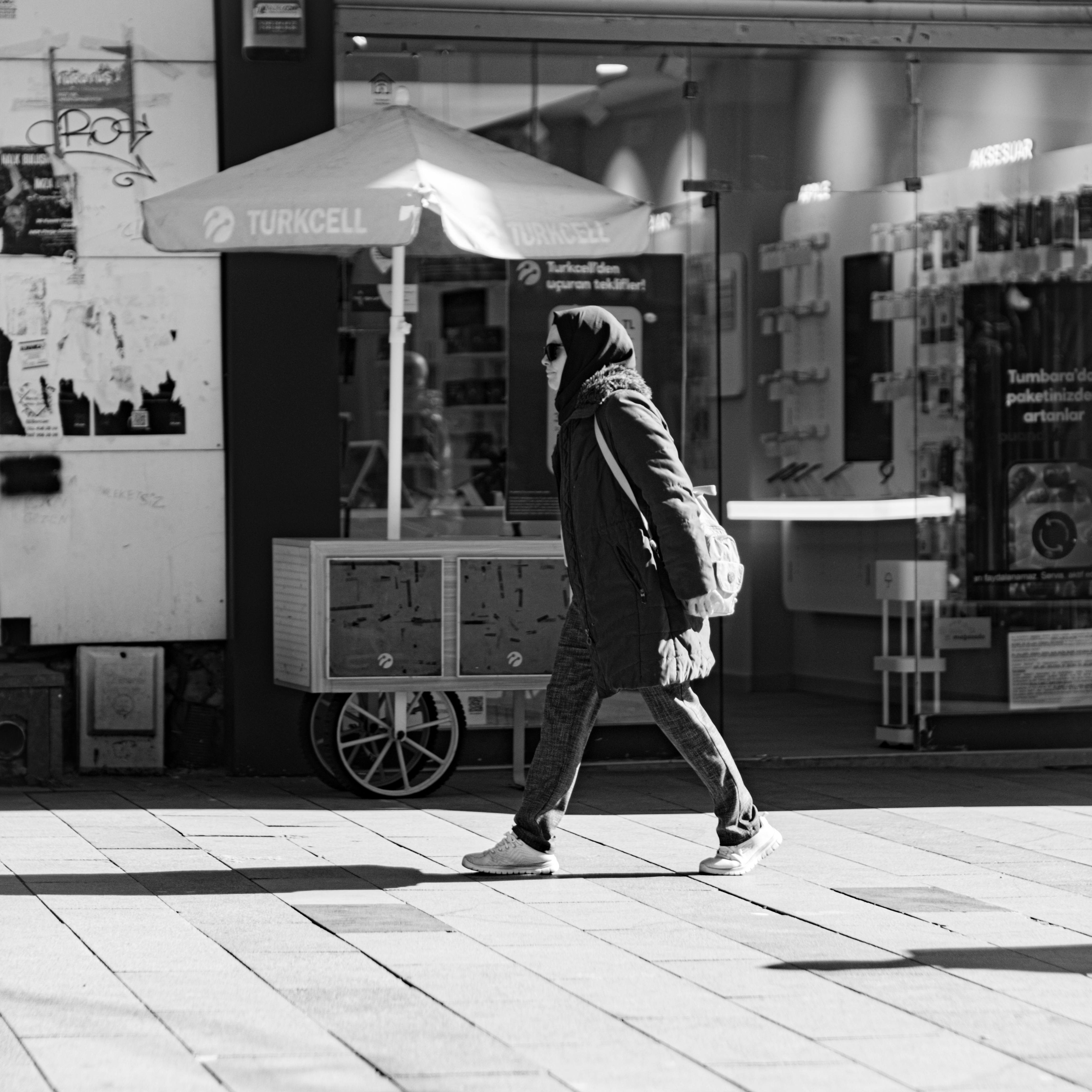 Free A woman in sunglasses and headscarf walks by a street market cart in a bustling urban environment. Stock Photo