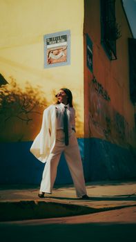 Fashionable woman in white suit posing on a sunny street corner with vibrant graffiti walls.