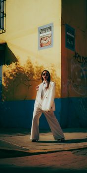Stylish woman in white suit poses confidently on a vibrant city street corner with graffiti.