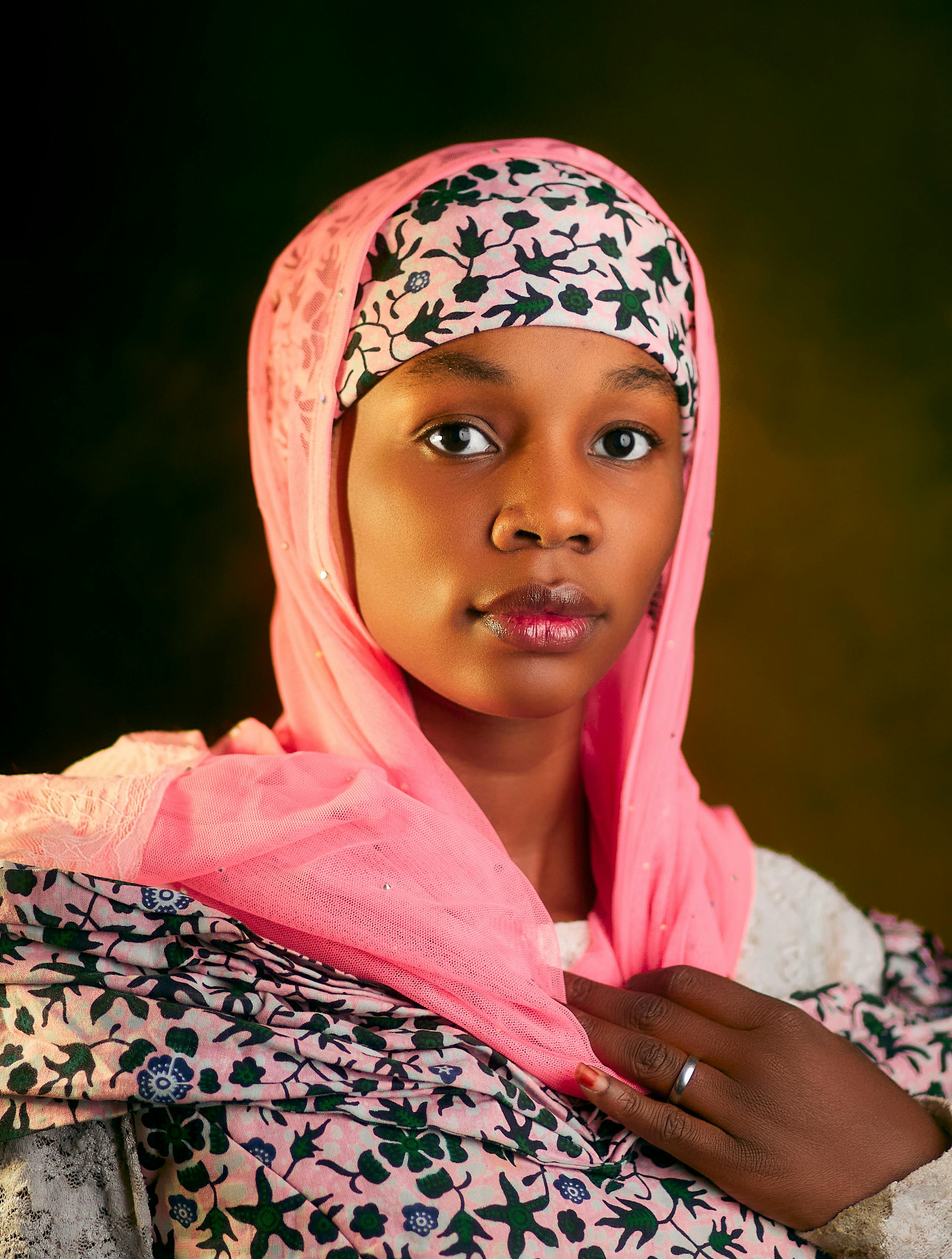 Elegant portrait of a woman wearing a traditional pink headscarf with floral patterns, against a dark moody background.