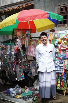 Street vendor smiling in front of a colorful toy stall under a rainbow umbrella on a vibrant day. - Fotografer Sidoarjo