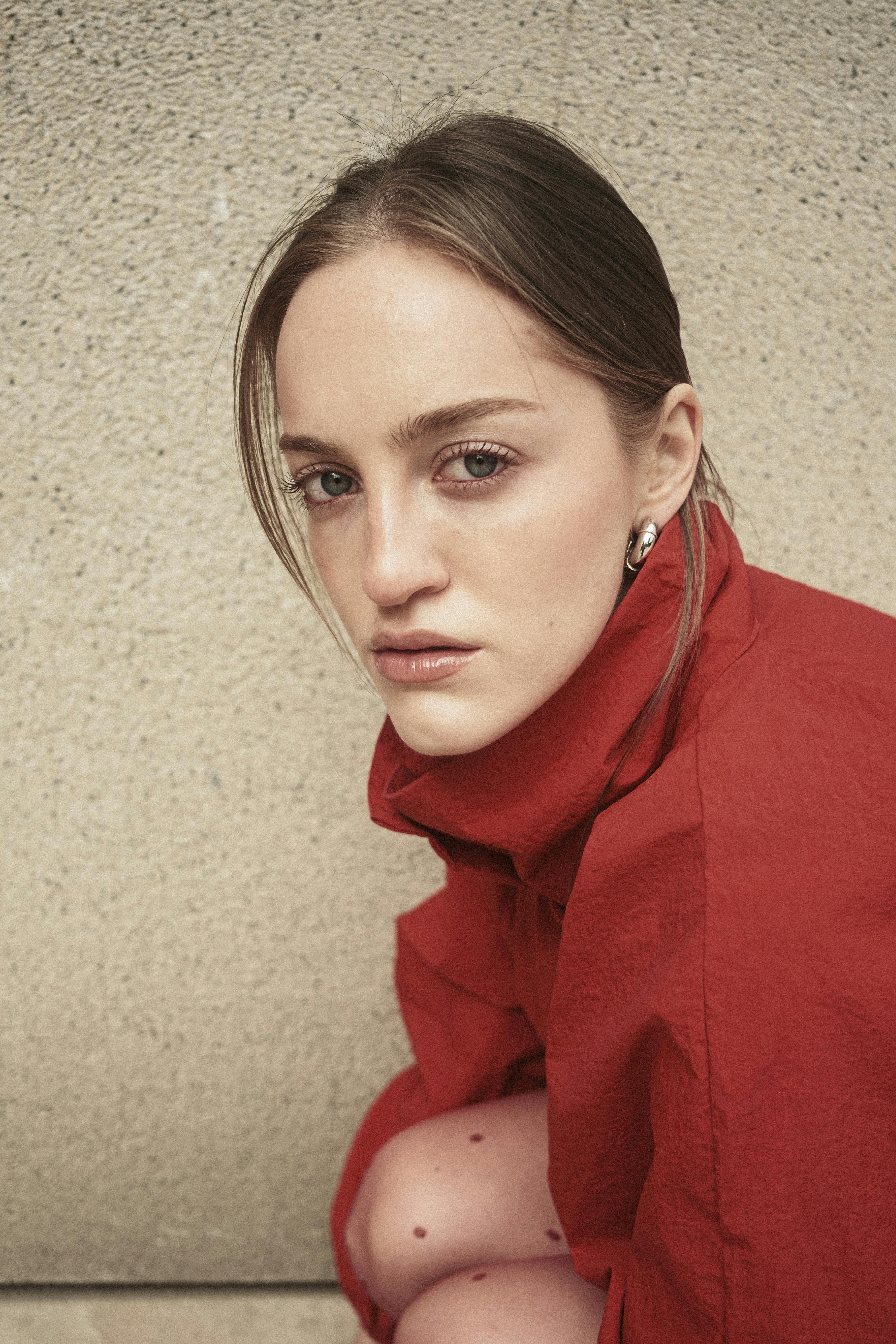 Elegant portrait of a woman in a red coat against a textured wall, showcasing modern fashion.