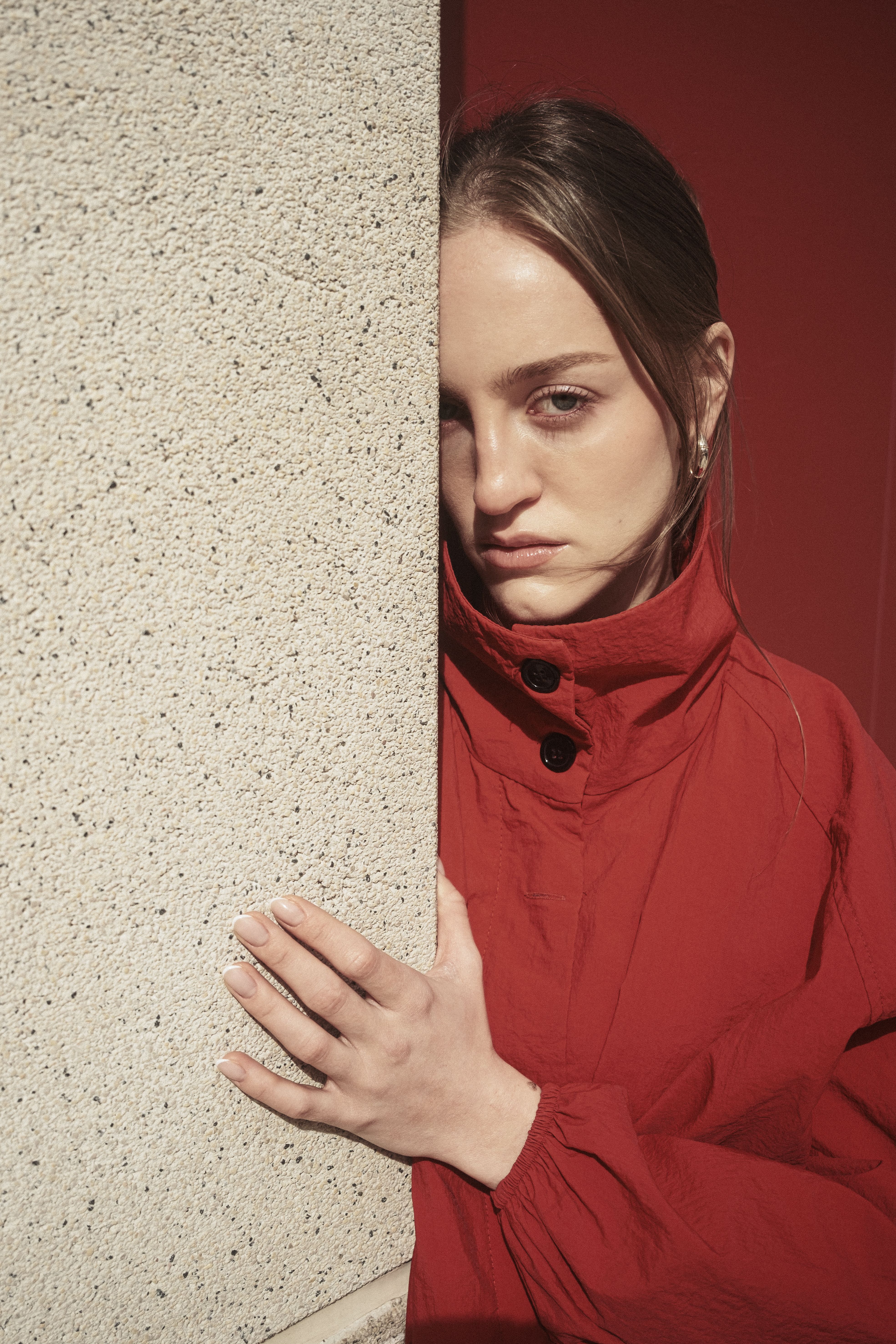 A thoughtful woman in a red coat leans against a textured wall, outdoors.