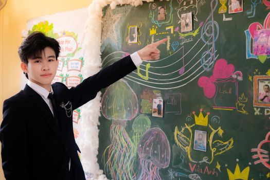 Young man in suit pointing at a colorful chalkboard with drawings and photos in a classroom setting.