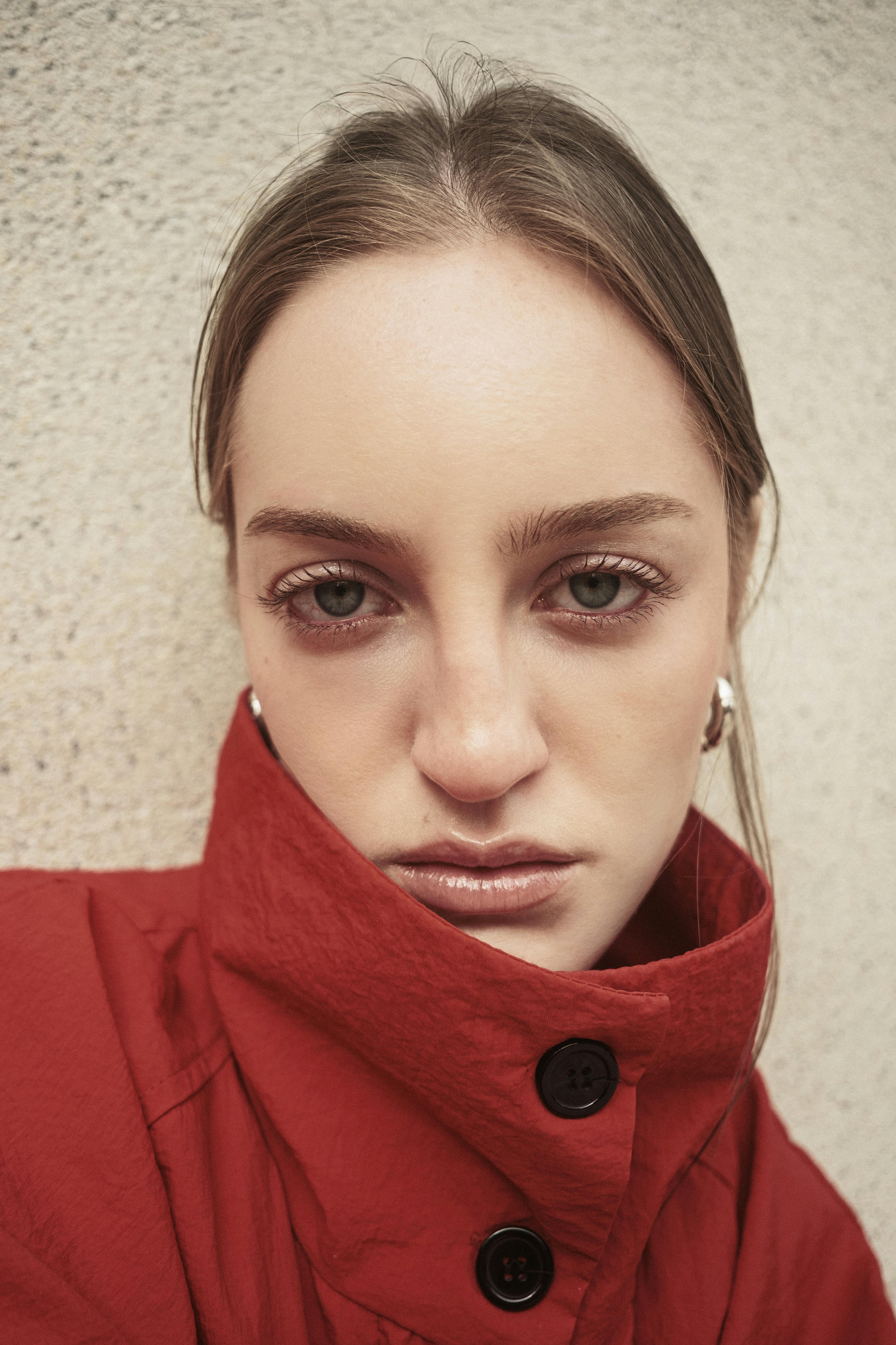 Close-up portrait of a woman in a red coat posing against a textured wall.