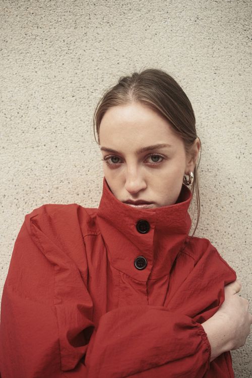 Free A young woman poses against a textured wall wearing a stylish red coat. Stock Photo