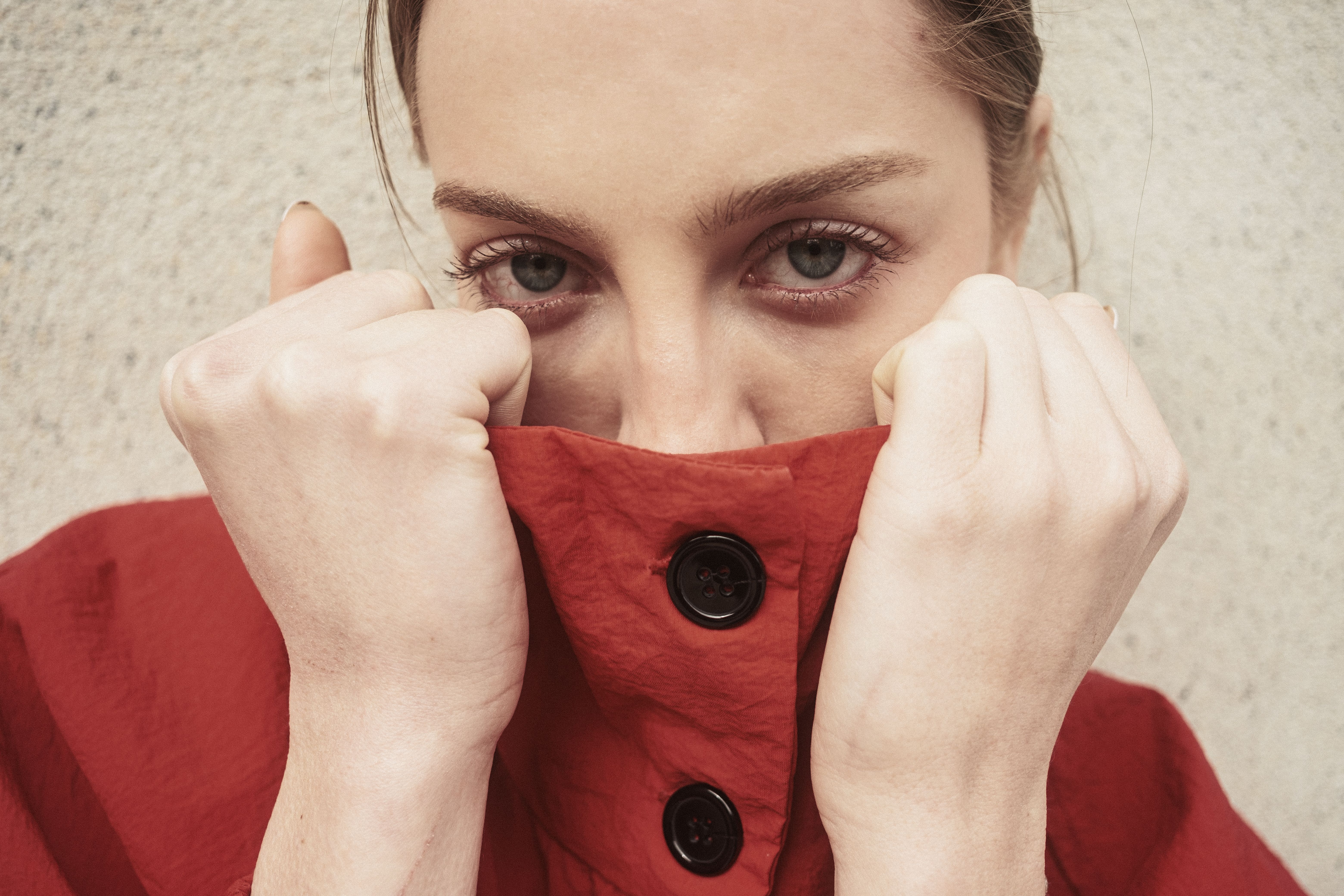 Close-up of a woman in a red coat showing a mysterious expression with a wall background.