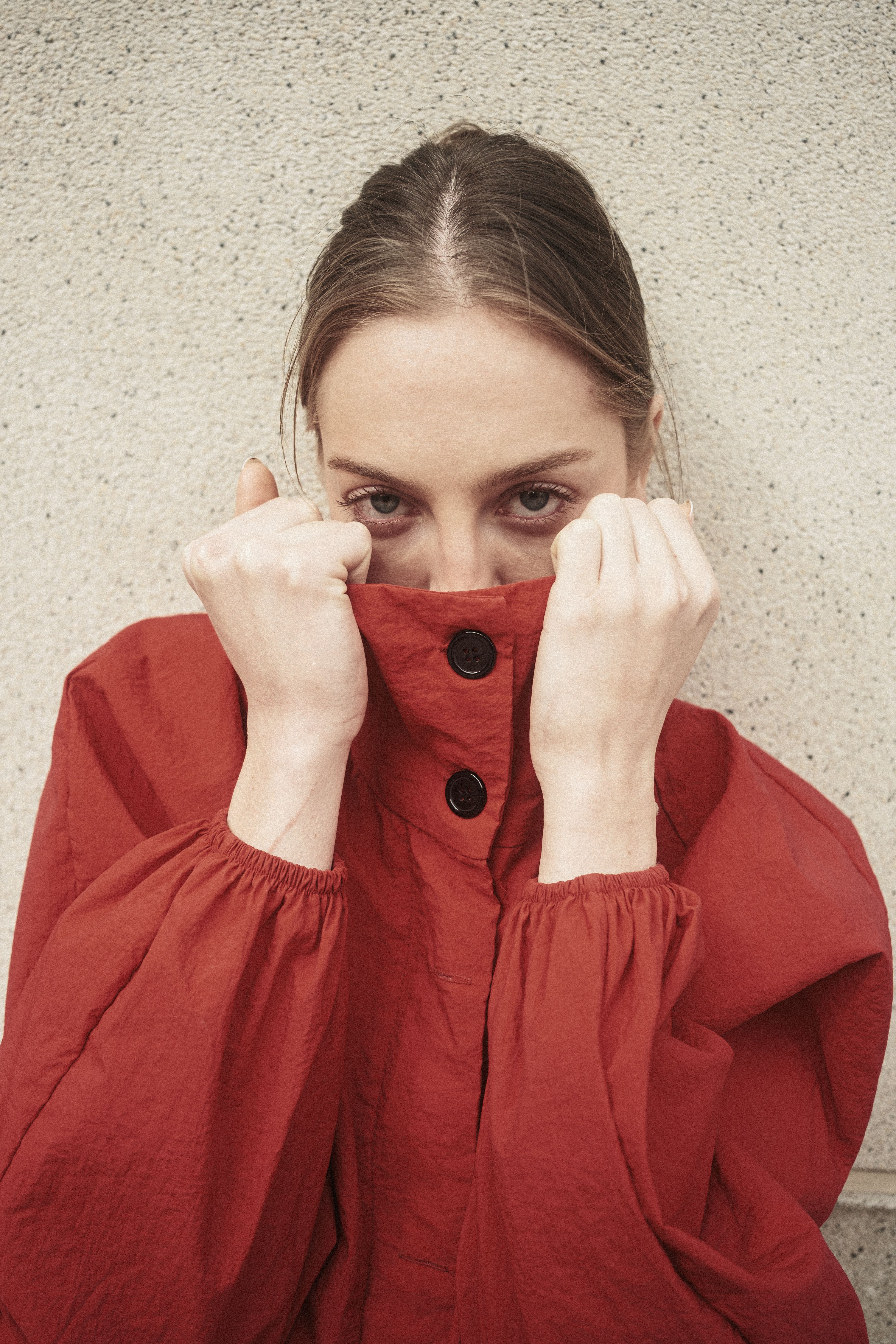 A woman playfully hides her face in a red jacket against a textured wall.