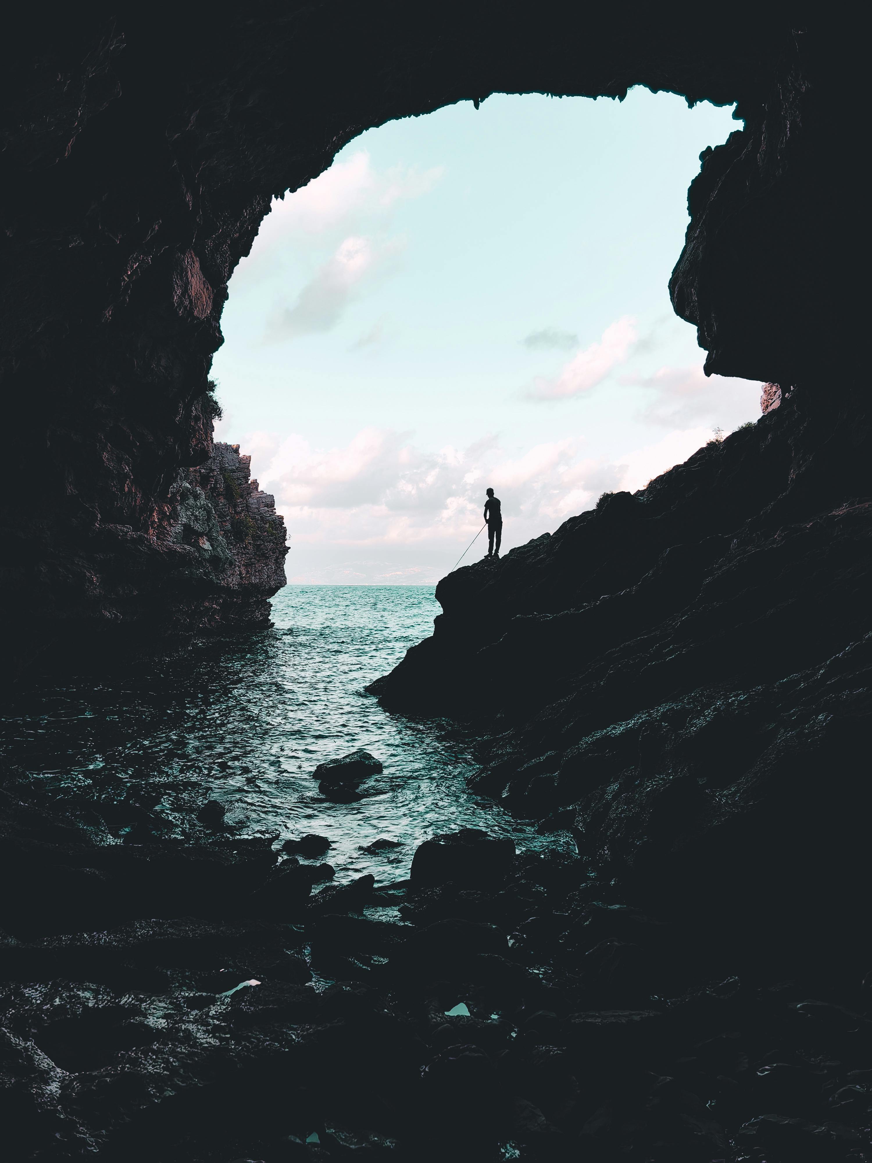 Free Dramatic silhouette of a person standing in a cave opening overlooking the ocean. Stock Photo