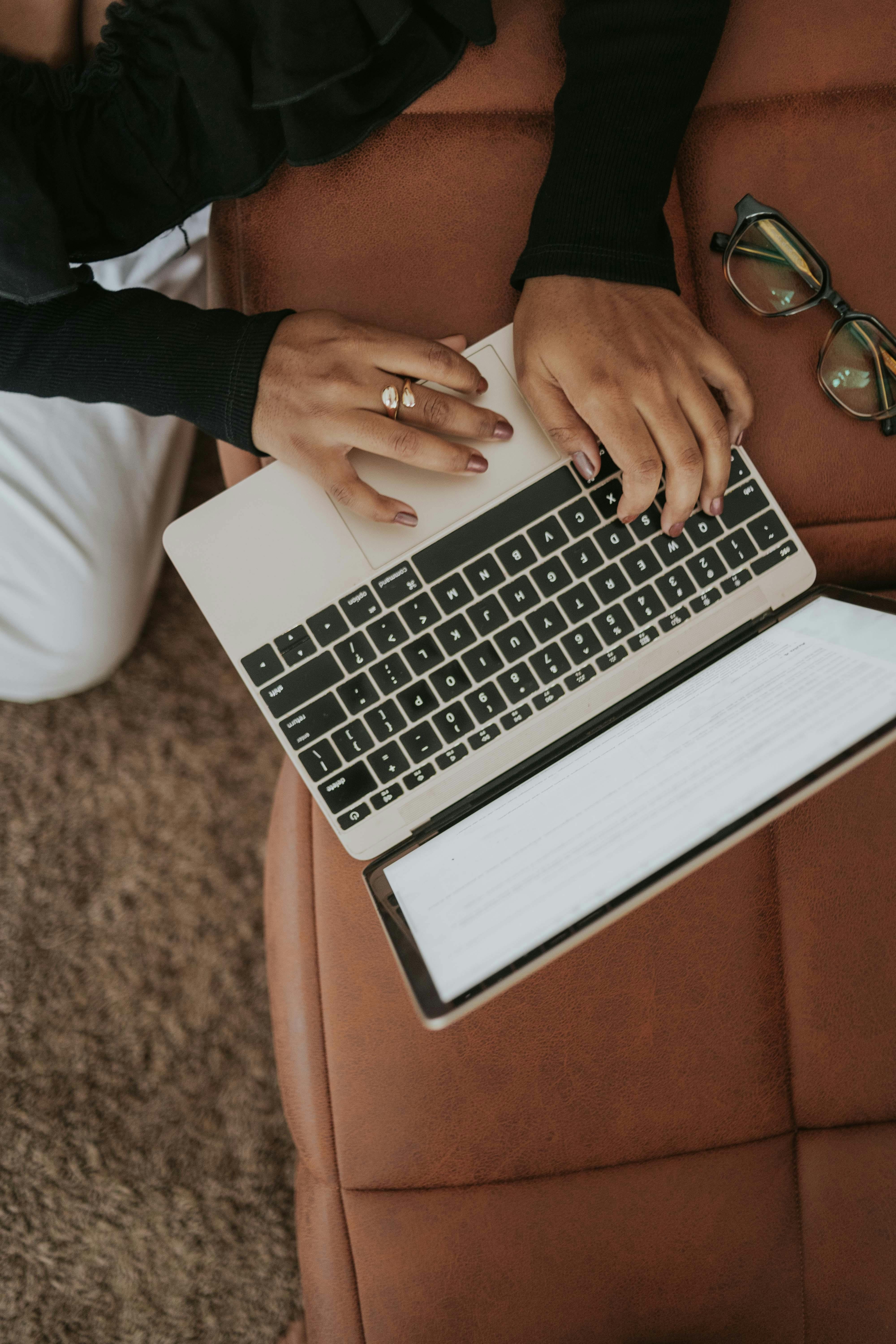 A woman wearing a ring types on a laptop on a brown couch with eyeglasses beside her.