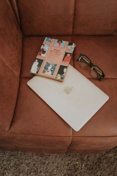 Modern flat lay featuring a stylish laptop, a colorful book, and glasses on a cozy brown chair.