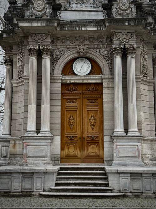 Free Elegant neoclassical building facade featuring ornate columns, a large wooden door, and a round clock above. Stock Photo