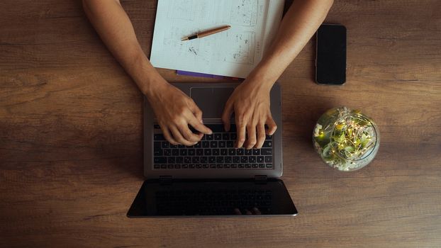 A person typing on a laptop from above, surrounded by papers and a smartphone on a wooden table.