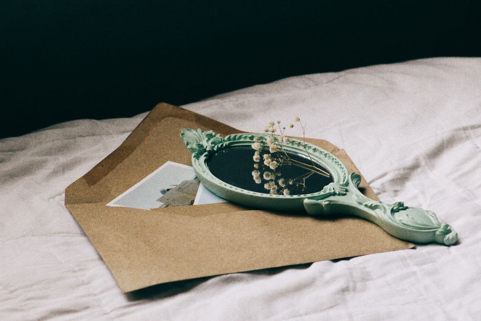 A vintage hand mirror and dried flowers lie atop a large rustic envelope on a soft surface.