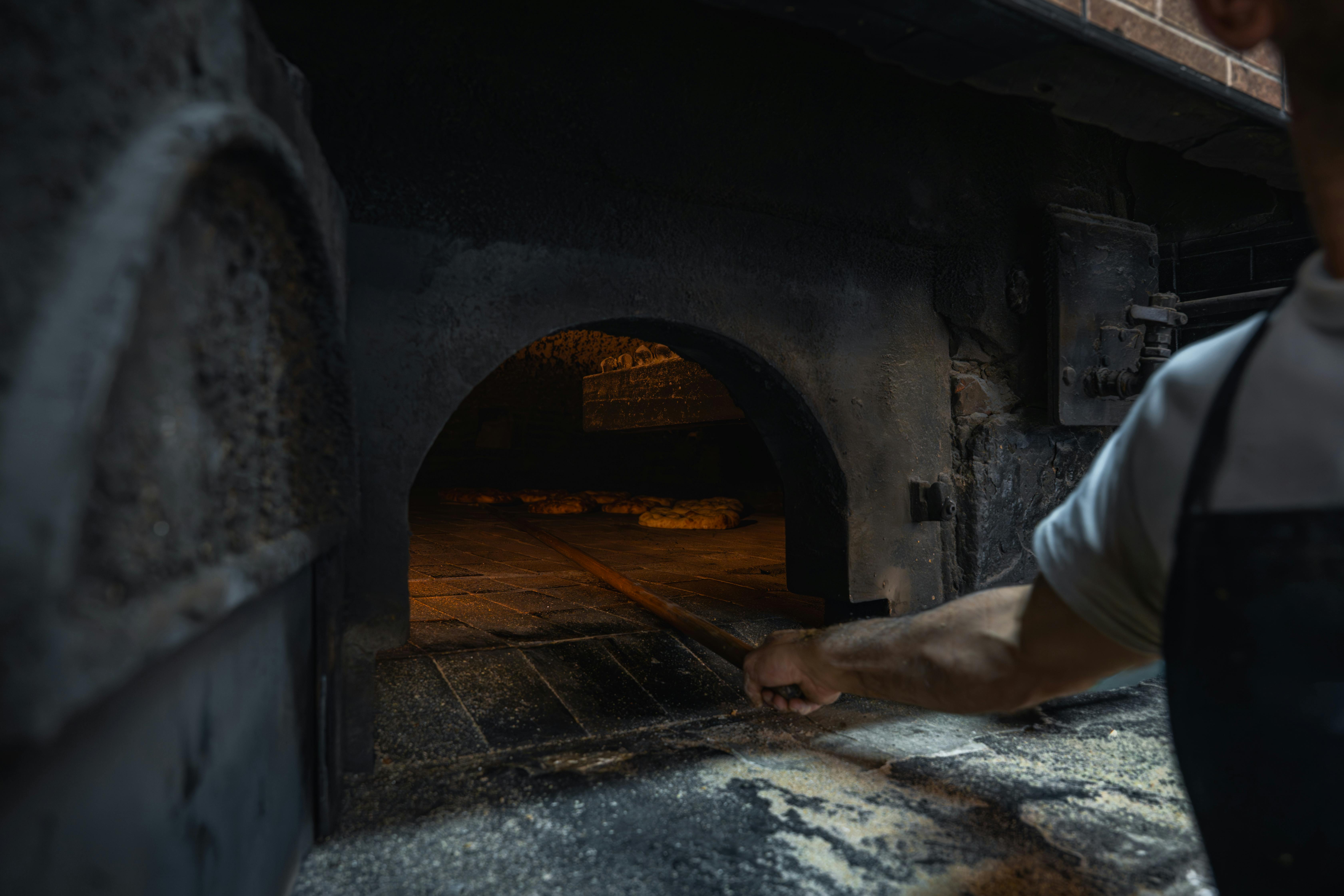 Baker using traditional oven for fresh bread preparation in a rustic setting.