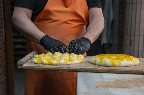 Free Chef in orange apron prepares cheesy bread with black gloves in a professional kitchen. Stock Photo