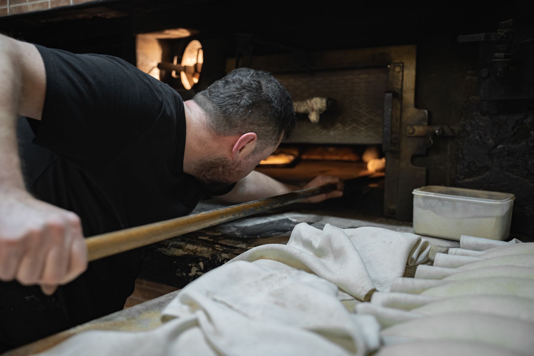 Panadero inclinado sobre la boca del horno, metiendo la pala; delante, piezas formadas entre lienzos
